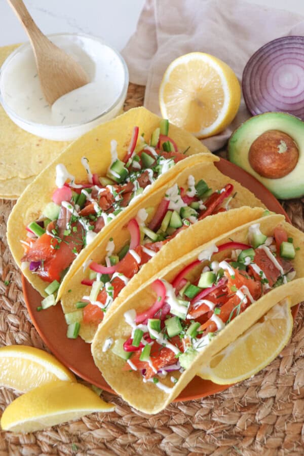 Corn tortillas filled with smoked salmon, diced cucumber, coleslaw and pickled red onions, garnished with dill and chives and topped with cilantro lime crema. Half an avocado, slices of lemon and a red onion and a white prep bowl with crema and a wooden spoon all on a woven mat.
