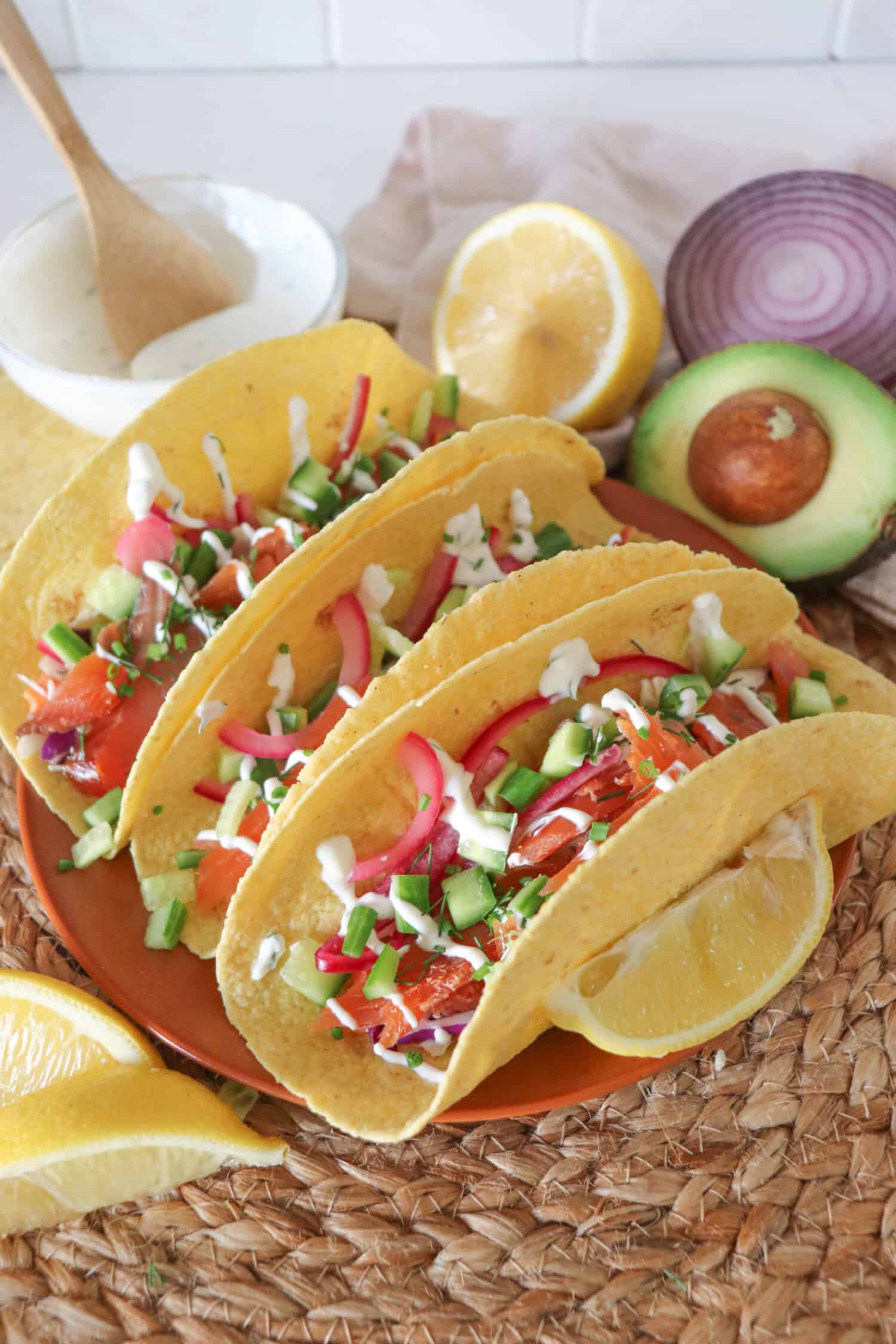 Corn tortillas filled with smoked salmon, diced cucumber, coleslaw and pickled red onions, garnished with dill and chives and topped with cilantro lime crema. Half an avocado, slices of lemon and a red onion and a white prep bowl with crema and a wooden spoon all on a woven mat.