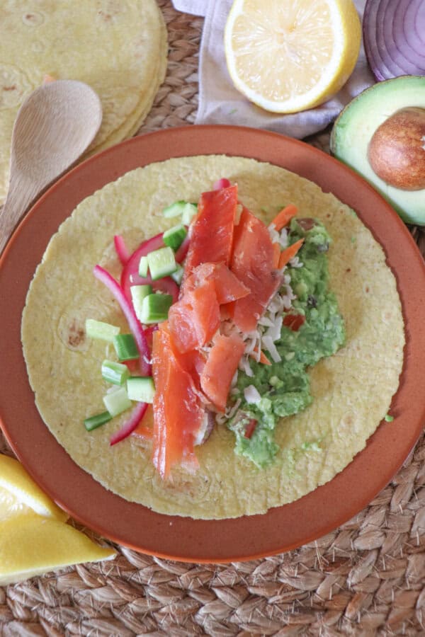 Open faced taco with smoked salmon, diced cucumbers, coleslaw, pickled red onion, guacamole on an orange plate. Half an avocado a wooden spoon and half a lemon also.