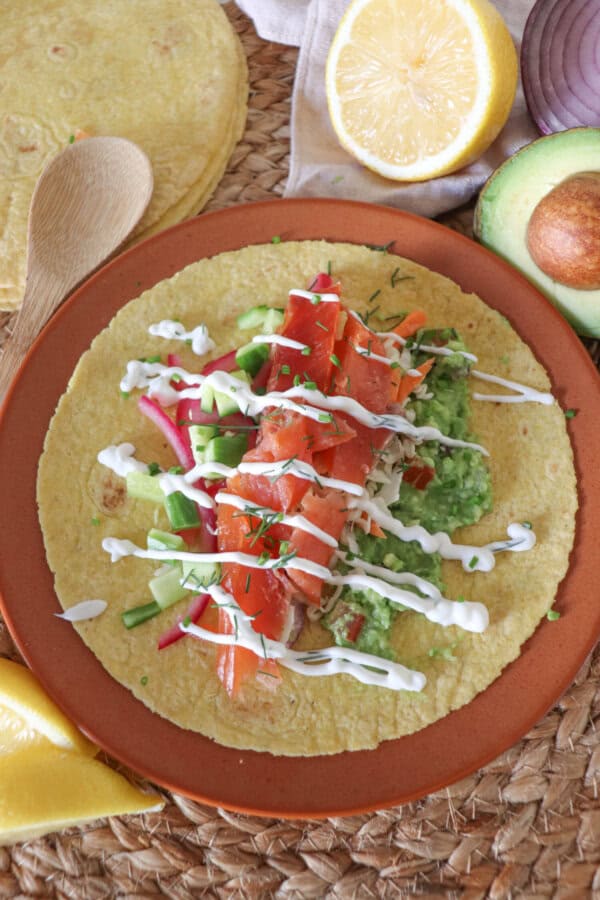 Open faced taco with smoked salmon, diced cucumbers, pickled red onion, guacamole and garnished with dill and chives and cilantro lime crema on an orange plate.

