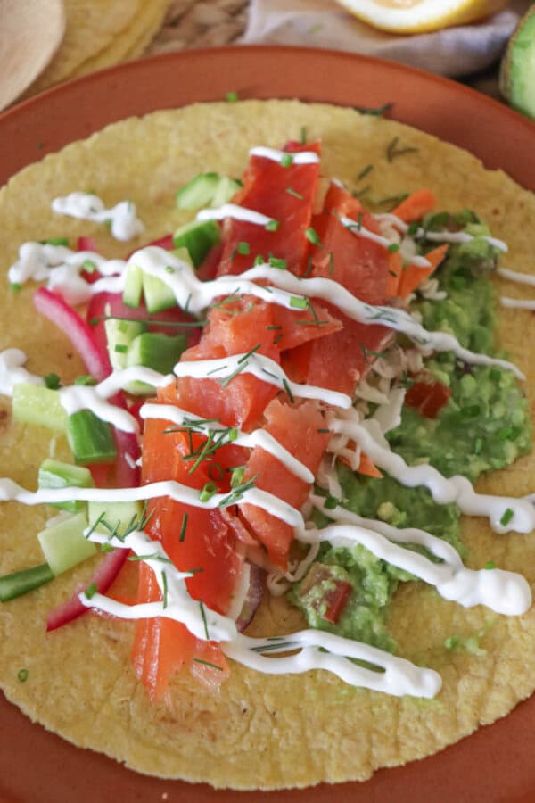 Open faced taco with smoked salmon, diced cucumbers, pickled red onion, guacamole and garnished with dill and chives and cilantro lime crema on an orange plate.