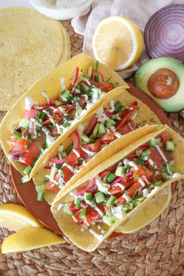 Corn tortillas filled with smoked salmon, diced cucumber, coleslaw and pickled red onions, garnished with dill and chives and topped with cilantro lime crema. Half an avocado, slices of lemon and a red onion and a white prep bowl with crema and a wooden spoon all on a woven mat.
