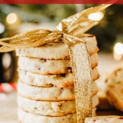 A stack of cranberry pistachio shortbread cookies tied with a gold ribbon is displayed. There are a few broken cookie pieces in the foreground. Text at the bottom reads homemadeheather.com.