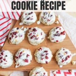 Overhead view of several cookies topped with white drizzle and dried cranberries on a wooden board. A red-striped cloth and additional cookies are in the background. Text reads: Cranberry Bliss Cookie Recipe and Homemade Heather.