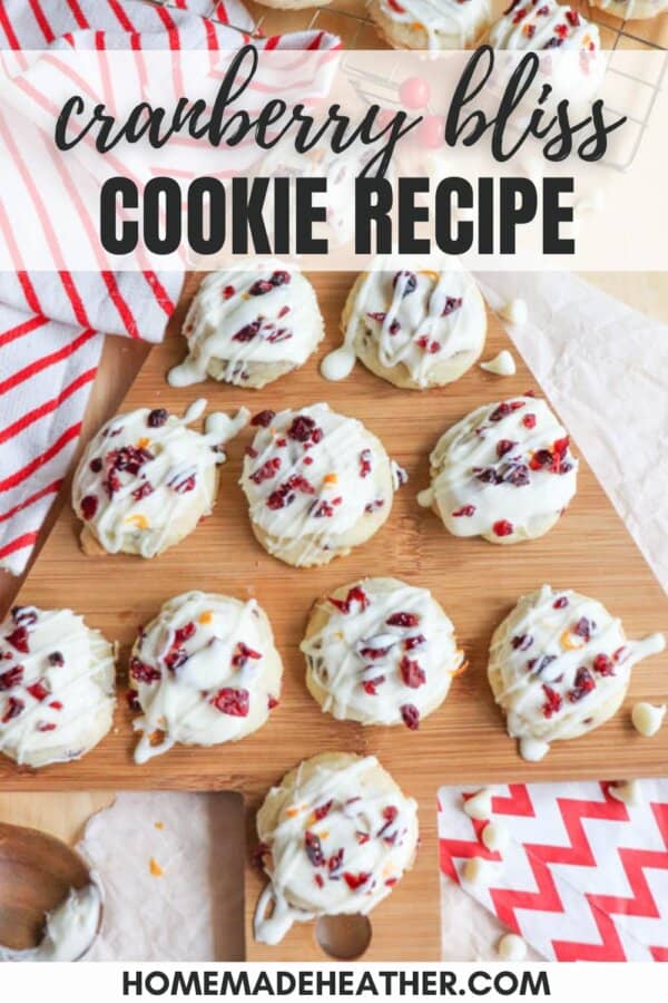 Overhead view of several cookies topped with white drizzle and dried cranberries on a wooden board. A red-striped cloth and additional cookies are in the background. Text reads: Cranberry Bliss Cookie Recipe and Homemade Heather.
