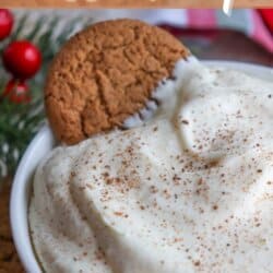 Close-up of a bowl filled with creamy eggnog dessert dip, topped with cinnamon. A gingerbread cookie is partially dipped into the mixture. Some festive decorations, including red berries and greenery, are visible in the background. Text reads: Eggnog dessert dip.