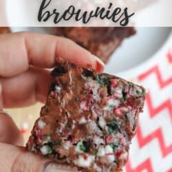 A close-up of a hand holding a candy cane brownie topped with crushed peppermint pieces. In the background, a plate with more brownies and a red and white zigzag-patterned napkin are visible. Text on the image reads CANDY CANE brownies.
