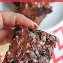 A hand holds a festive brownie with colorful candy cane pieces on top. A plate with more brownies is in the background, placed on a red and white chevron-patterned napkin. The text Candy Cane Brownies is at the top.