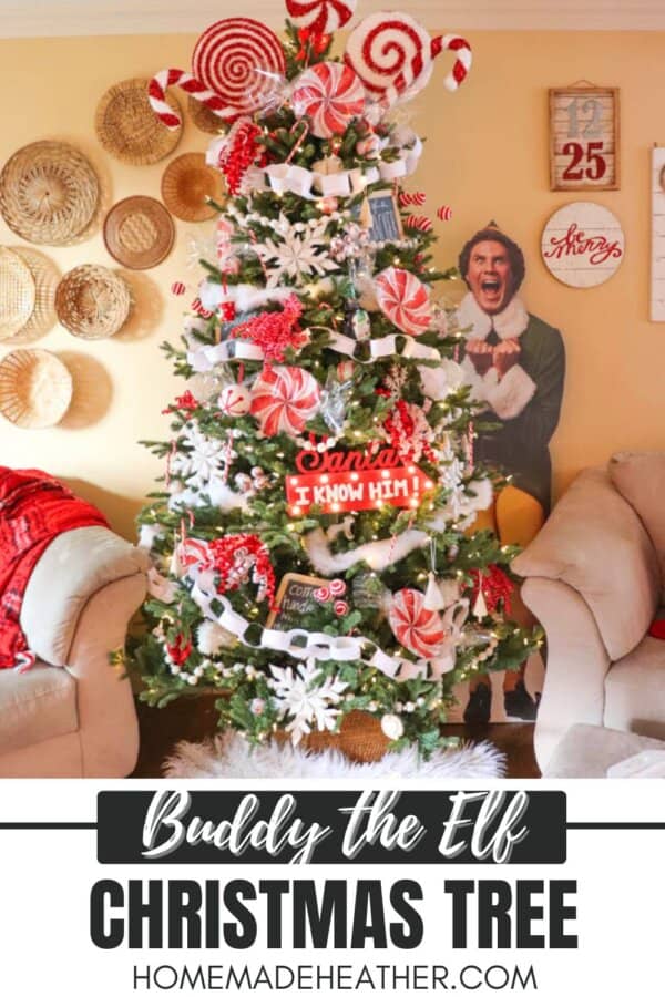 A Christmas tree decorated with red and white candy ornaments, snowflakes, and greenery. It features a cardboard cutout of Buddy the Elf next to it. The room has beige walls with baskets and a decorative sign.