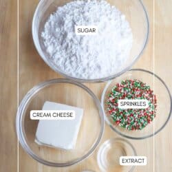 Top-down view of baking ingredients on a wooden surface: a bowl of powdered sugar, a bowl with a block of cream cheese, a bowl of colorful sprinkles, a small bowl of liquid extract, and a small cup of milk.