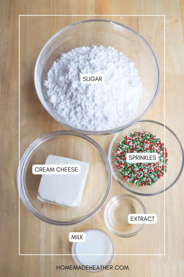 Top-down view of baking ingredients on a wooden surface: a bowl of powdered sugar, a bowl with a block of cream cheese, a bowl of colorful sprinkles, a small bowl of liquid extract, and a small cup of milk.