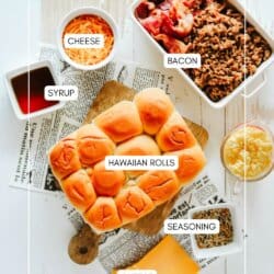 Top view of a flatlay featuring ingredients for sliders: Hawaiian rolls, grated cheese, pieces of bacon in a dish, syrup in a cup, and seasonings next to a small bowl. Slices of cheese and a newspaper are also visible beneath the rolls.