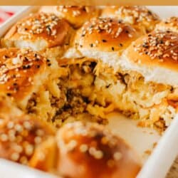 A baking dish filled with breakfast sliders made of soft buns topped with sesame seeds. The sliders are stuffed with scrambled eggs, cheese, and sausage, with one slider partially pulled out. Text on image reads breakfast sliders and homemadeheather.com.