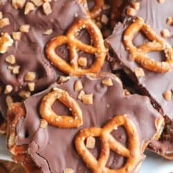Close-up of chocolate pretzel toffee bark pieces stacked on a plate. The bark is topped with chocolate, pretzel twists, and bits of toffee. Red and white candy canes are visible in the background. Text reads Chocolate Pretzel Toffee Bark and Homemade Heather.