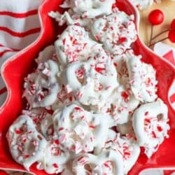 A festive red plate piled with white chocolate peppermint pretzels, sprinkled with crushed peppermint candy. The plate is shaped like a Christmas tree and is surrounded by red berries and a striped red and white cloth.