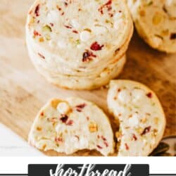 A stack of cranberry pistachio shortbread cookies on a wooden surface. The cookies are speckled with visible pieces of cranberries and pistachios. Text at the bottom reads shortbread cranberry pistachio homemakeheather.com.