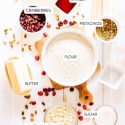 A flat lay of baking ingredients on a white wooden surface, including bowls of flour, cranberries, pistachios, vanilla, white chocolate, and sugar, with sticks of butter. Labels identify each ingredient.