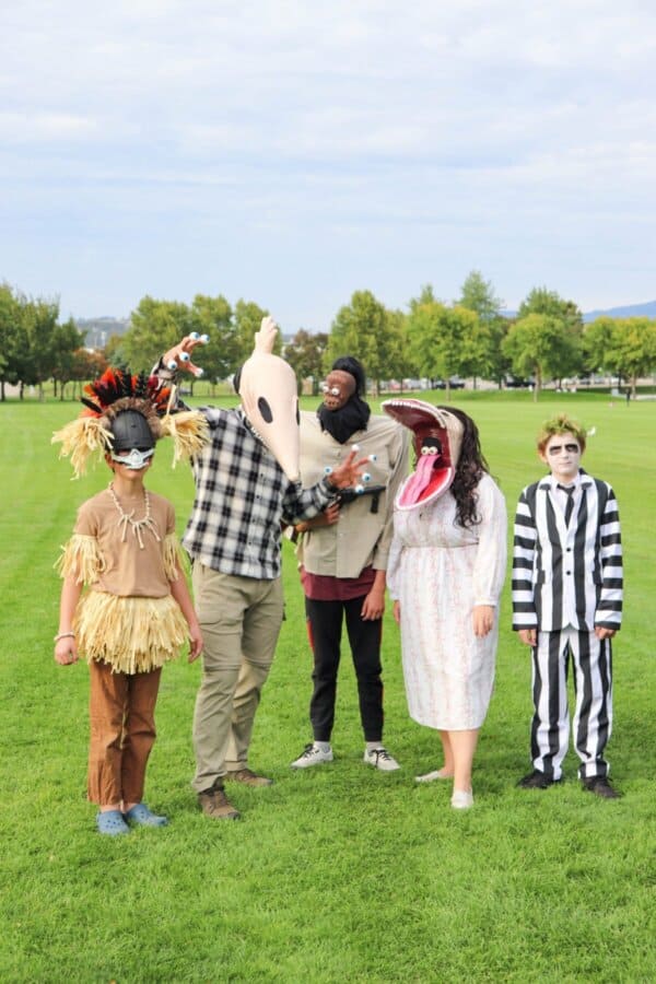 Five people stand on grass in homemade costumes. Their outfits feature a skull mask, jungle attire, a large mouth headpiece, and a black-and-white striped suit. Heather trees and a cloudy sky provide the perfect eerie backdrop.