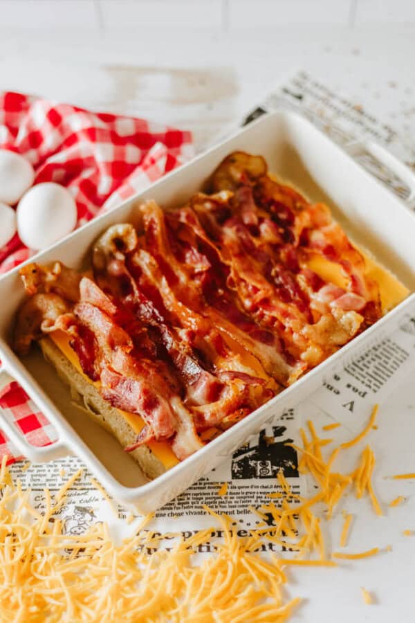 A white baking dish filled with baked bacon and melted cheese. Surrounding the dish are scattered shredded cheese, a red and white checkered cloth, and three white eggs against a newspaper-textured background.