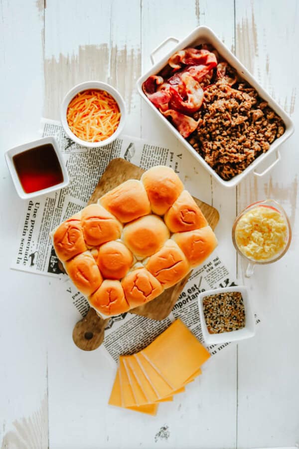 A flat lay of sandwich ingredients on a white wooden table. Includes a tray of pulled meat, a pan of buns, cheese slices, grated cheese, pineapple, bacon, syrup, and seasoning in bowls, with newspaper beneath some items.