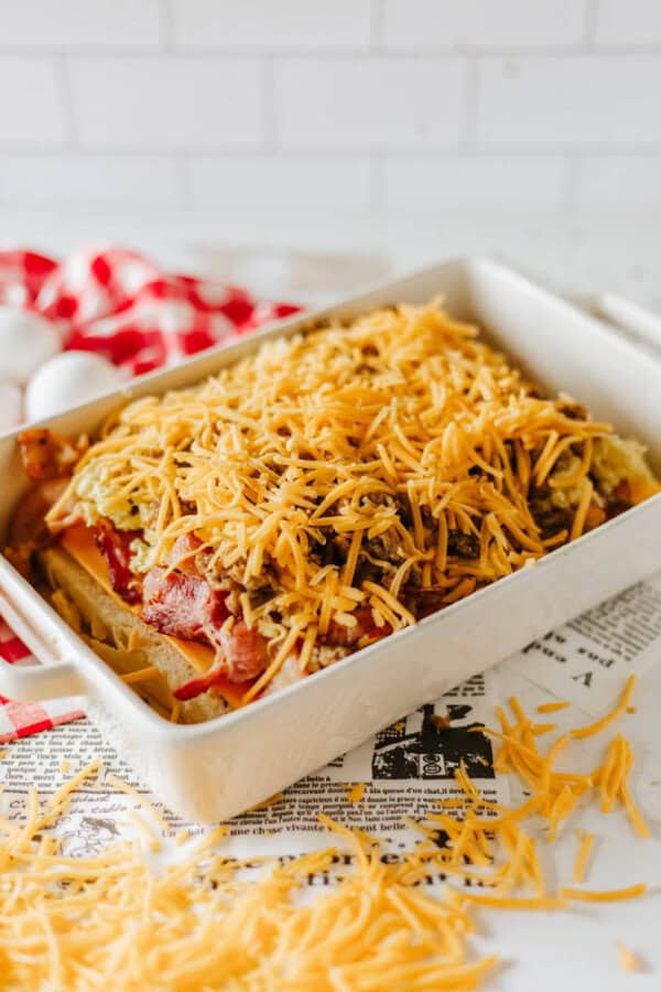 A baking dish filled with a layered casserole topped with shredded cheese, ready for baking. The dish rests on a newspaper, with scattered cheese on the surface and a red and white checkered napkin in the background.