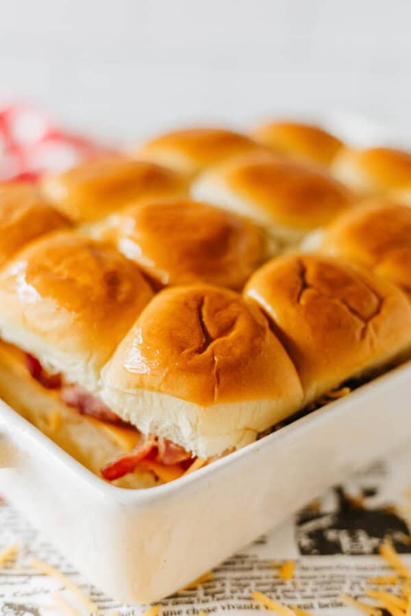 A close-up of golden-brown slider buns arranged in a baking dish. The sliders are filled with layers of cheese and bacon, sitting on a bed of crumpled newspaper. The setting is bright and inviting.