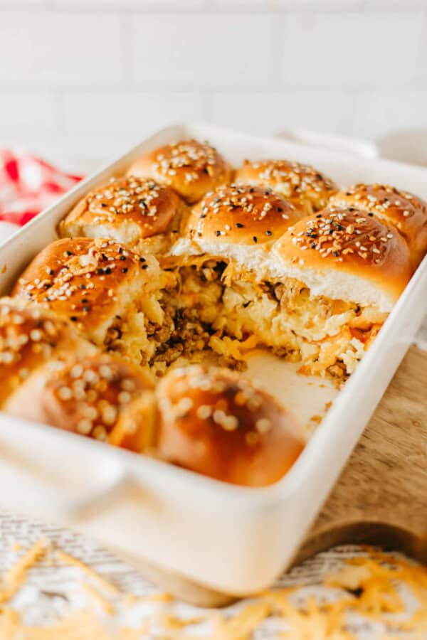 A baking dish filled with golden brown burger sliders topped with sesame seeds. One slider in the center is missing, revealing the filling of cooked ground meat and melted cheese. The background includes a red and white checkered cloth.