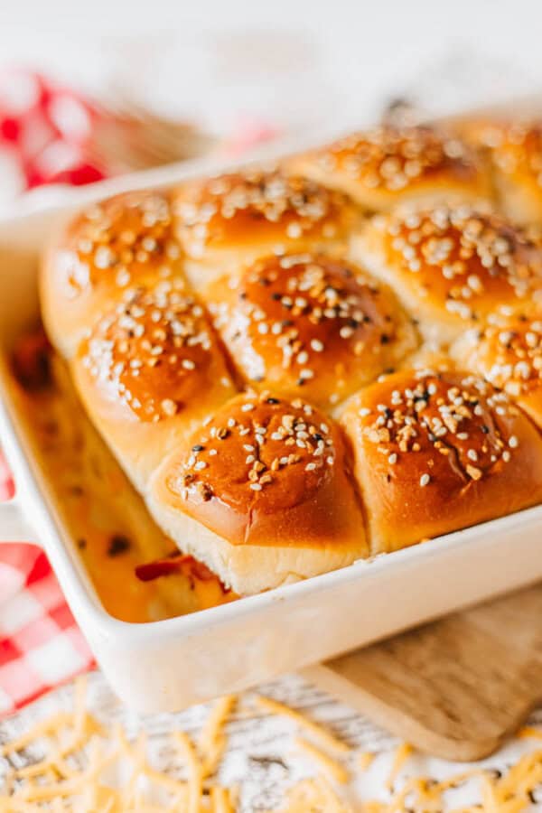 A baking dish filled with golden-brown slider buns topped with sesame seeds. The buns are closely packed and appear freshly baked, sitting on a checkered cloth with scattered shredded cheese around.