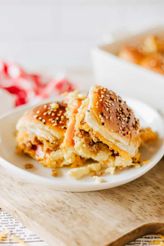 A close-up of two cheesy beef sliders on a white plate. The sliders are topped with sesame seeds and placed on a wooden surface. The background features a red and white checkered cloth and a hint of a baking dish.