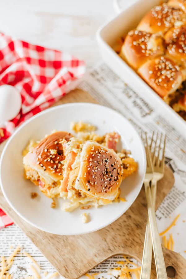 A plate of fluffy, pulled-apart dinner rolls topped with sesame seeds sits on a wooden board. The rolls are beside a fork and knife, with a baking dish of similar rolls and a red and white checkered cloth in the background.
