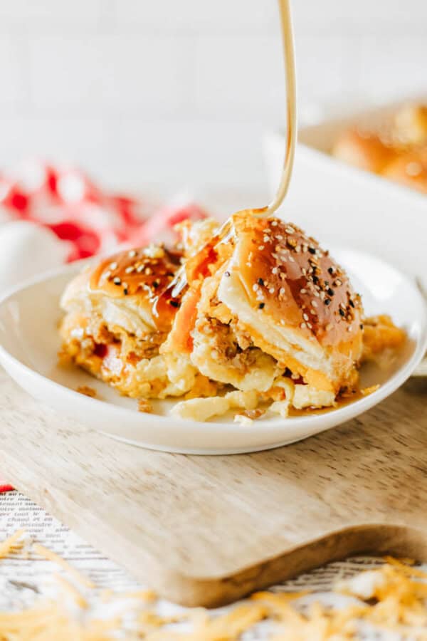 A savory slider topped with sesame seeds is being drizzled with honey on a white plate. The slider has a golden-brown bun and is filled with a cheesy, meaty mixture. Its placed on a wooden cutting board, with a checkered cloth in the background.