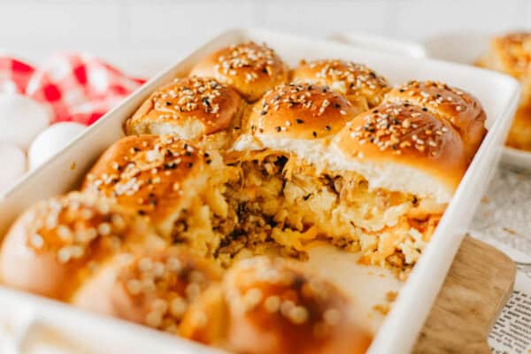 A baking dish filled with golden-brown, freshly baked rolls. The rolls are topped with sesame and poppy seeds and are filled with a mix of scrambled eggs and sausage, with some removed to show the filling.
