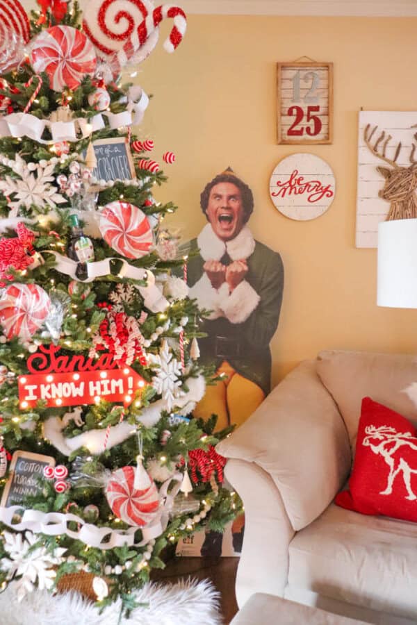 A festive living room with a Christmas tree adorned with red and white decorations, including candy-shaped ornaments. A cutout of a person in an elf costume is behind the tree. Holiday-themed signs are on the yellow wall, near a beige couch.