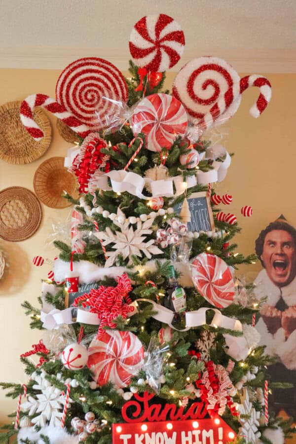 A Christmas tree decorated with candy cane and peppermint swirls, white ribbons, and twinkling lights. A Santa, I know him! sign is at the bottom. A poster of an excited character is partially visible in the background.