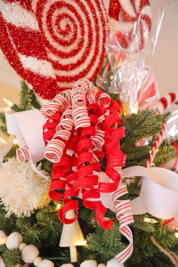 Close-up of a Christmas tree decorated with red and white swirl lollipops, curly ribbons, and paper chains. The tree is adorned with warm white lights and features festive, candy-themed ornaments.