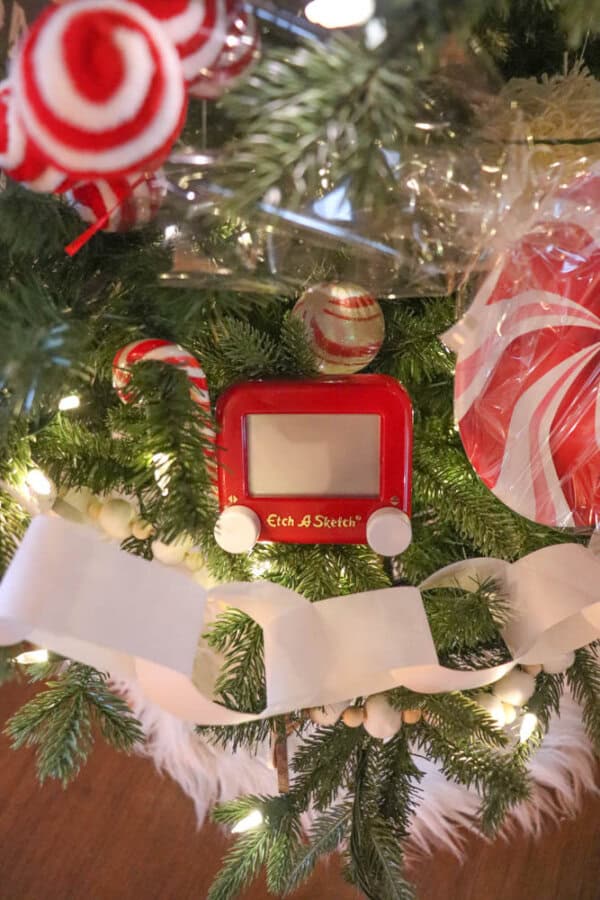 A decorated Christmas tree with a mini red Etch A Sketch ornament, candy cane, and swirled lollipop ornament. White paper garland and string lights are intertwined with green branches, creating a festive holiday display.