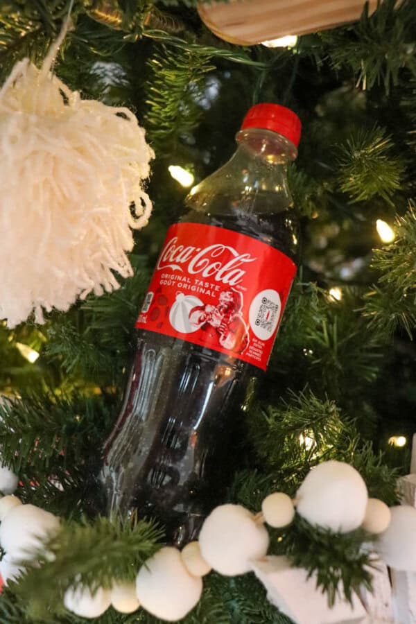 A Coca-Cola bottle nestled among branches of a decorated Christmas tree, adorned with white lights, a knitted white ornament, and round white decorations, creating a festive atmosphere.