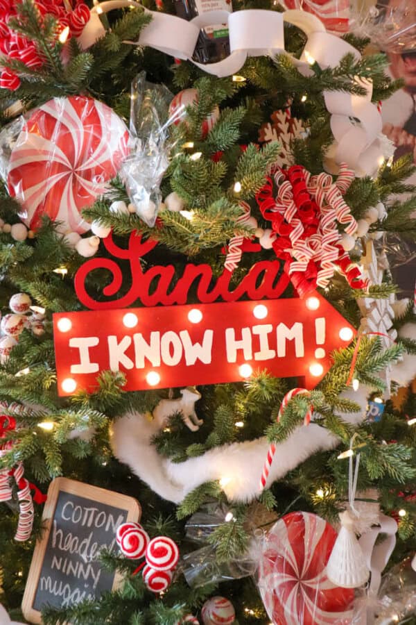 A festive Christmas tree decorated with red and white striped ornaments, candy canes, and ribbon. A sign on the tree reads Santa, I know him! in bold red and white letters. The tree is lit with warm white lights.