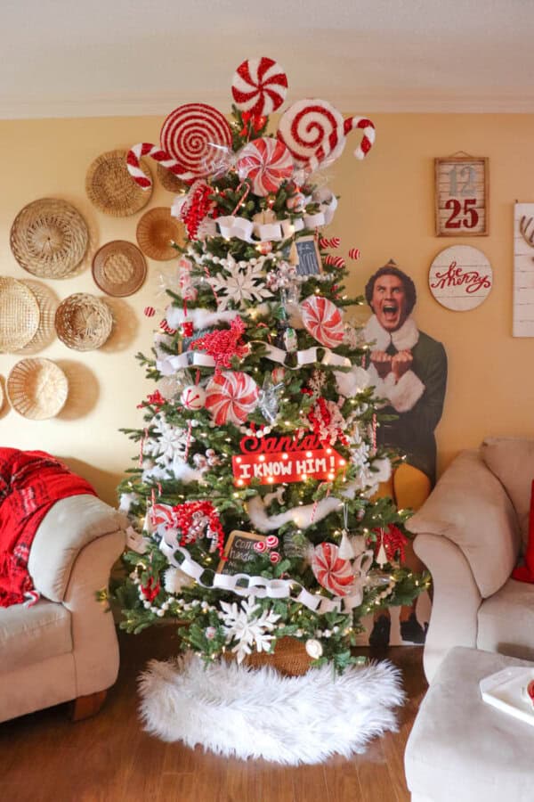 Star Wars Chewbacca themed Christmas tree A festive Christmas tree adorned with candy-themed ornaments, including peppermint swirls and candy canes. A cutout of a cheerful man in an elf costume stands beside it. Cozy living room with beige couches, red blankets, and wicker wall decor.