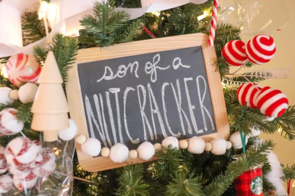 A Christmas tree decorated with white and red ornaments, candy canes, and a small wooden sign that reads Son of a Nutcracker in white chalky letters. The tree is adorned with lights and festive garlands.