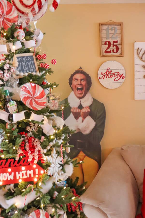 A festive room with a decorated Christmas tree featuring candy and ribbons. A life-sized cutout of a person in a green holiday costume is next to the tree. The wall has wooden holiday signs, including a calendar showing December 25.