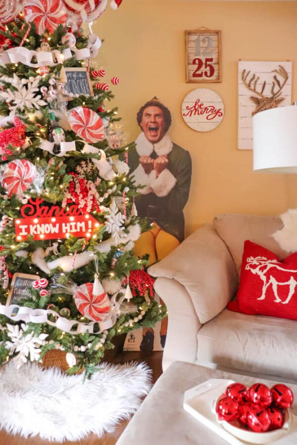 chewbacca christmas tree A festive living room features a decorated Christmas tree with red and white ornaments. A cardboard cutout of a man in an elf costume stands near the tree. A red pillow with a white reindeer design is on the sofa. A bowl of red ornaments is on the table.
