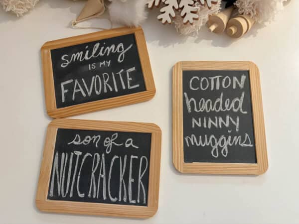 Three decorative chalkboards with playful phrases in white text: Smiling is my favorite, Son of a nutcracker, and Cotton-headed ninny muggins. They are surrounded by festive decorations.