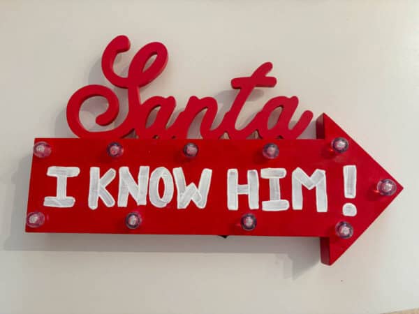 A decorative sign with a red arrow and lights reads Santa I KNOW HIM! in white text. The word Santa is stylized in cursive above the arrow. The background is plain.