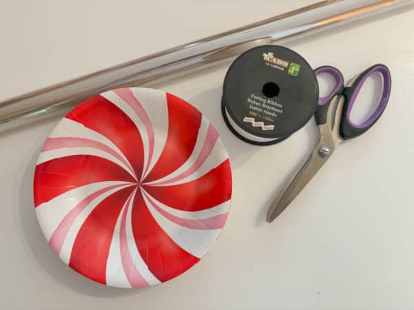 A red and white swirl-patterned paper plate, a pair of scissors with purple handles, a spool of black curling ribbon, and two long wooden dowels are arranged on a white surface.