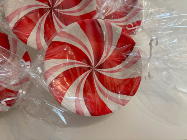 Image of several round plates with red and white spiral patterns, resembling peppermint candies, wrapped in clear plastic with white ribbons. The plates are arranged on a light surface.
