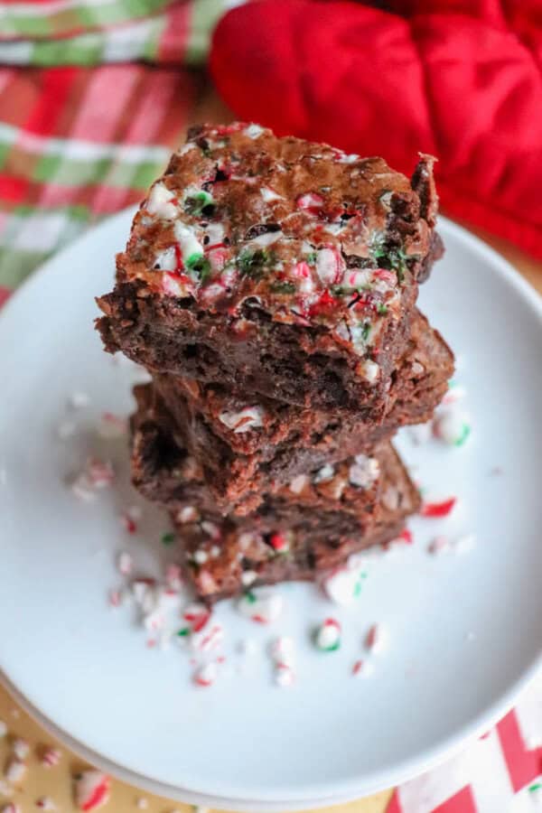 A stack of chocolate brownies topped with crushed peppermint candy on a white plate. The background has a red and green plaid fabric and a red oven mitt. Crushed candy pieces are scattered around the plate.