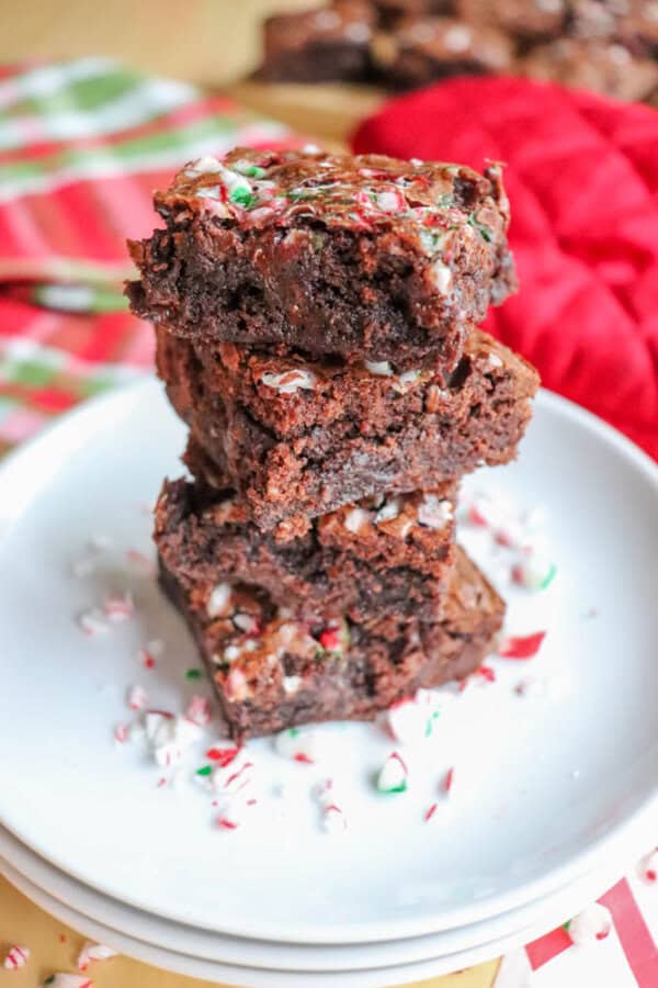 A stack of chocolate brownies topped with crushed candy canes sits on a white plate. Red and white striped fabric is in the background, adding a festive holiday touch. Crushed candy canes are scattered around the plate.