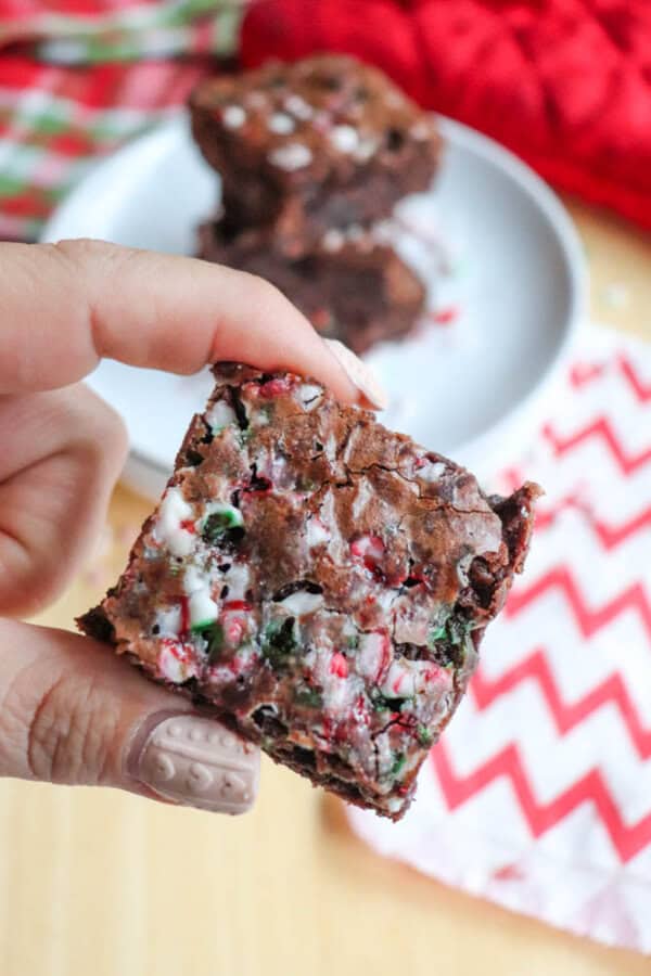 A hand holding a brownie piece topped with crushed candy canes. A blurry plate with more brownies is in the background. The tablecloths have red and white patterns.