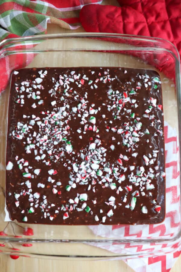 A glass baking dish with chocolate batter sprinkled with crushed peppermint candies. A red oven mitt and a chevron-patterned cloth are in the background.